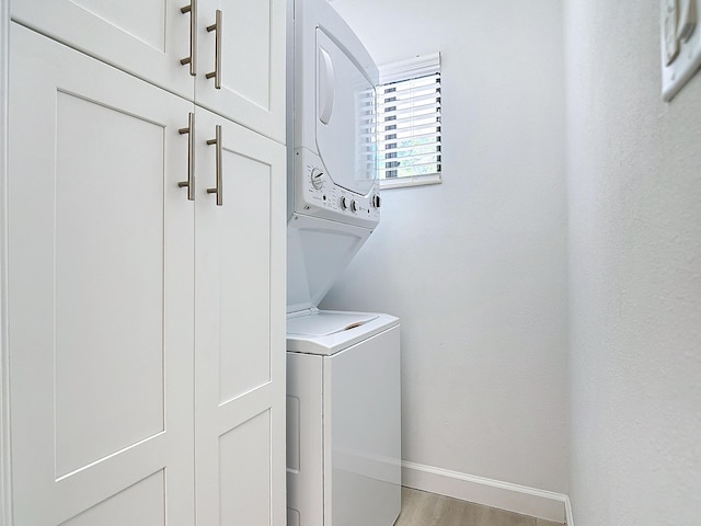 clothes washing area featuring light wood-type flooring, stacked washing maching and dryer, cabinet space, and baseboards