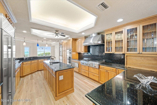 kitchen featuring wall chimney range hood, pendant lighting, a raised ceiling, a center island with sink, and appliances with stainless steel finishes