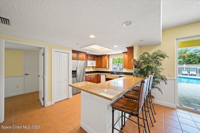 kitchen featuring light stone countertops, stainless steel refrigerator with ice dispenser, kitchen peninsula, a breakfast bar, and light tile patterned floors