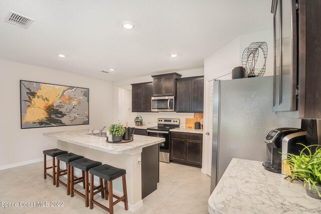 kitchen featuring dark brown cabinets, light tile patterned floors, a kitchen bar, appliances with stainless steel finishes, and a center island with sink