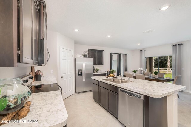 kitchen with light tile patterned flooring, dark brown cabinets, an island with sink, appliances with stainless steel finishes, and sink