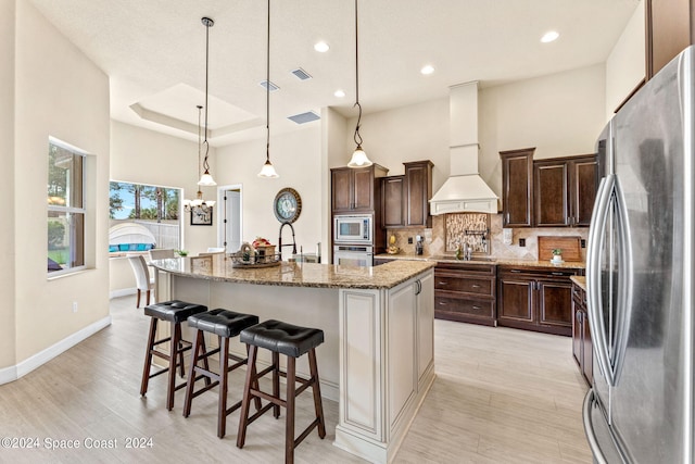 kitchen with pendant lighting, premium range hood, a center island with sink, stainless steel appliances, and light wood-type flooring