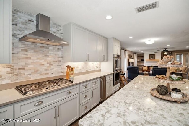 kitchen with ceiling fan, light stone countertops, wall chimney exhaust hood, backsplash, and appliances with stainless steel finishes