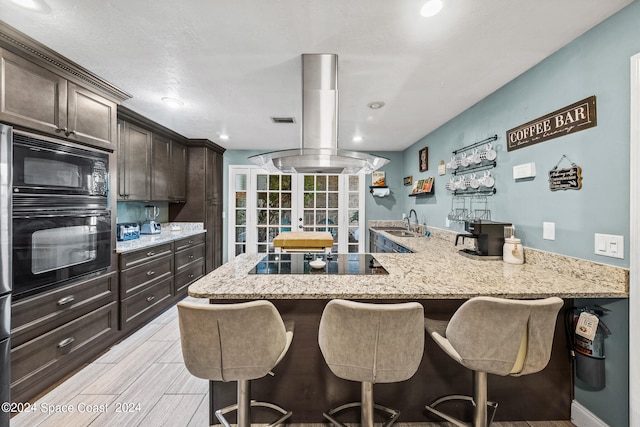 kitchen with a kitchen bar, dark brown cabinets, light stone counters, black appliances, and island exhaust hood