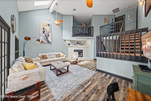 living room featuring ceiling fan, a skylight, a tiled fireplace, hardwood / wood-style flooring, and beamed ceiling