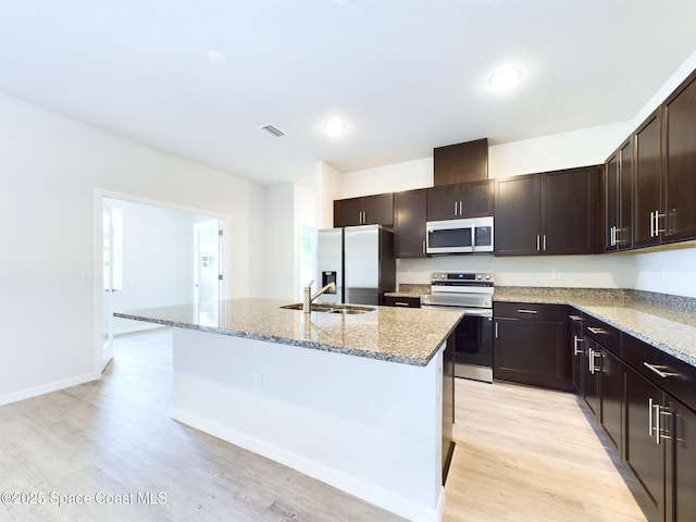 kitchen with sink, stainless steel appliances, light stone counters, a center island with sink, and dark brown cabinetry