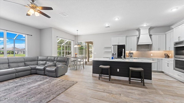 kitchen with white cabinets, a kitchen island with sink, decorative light fixtures, custom range hood, and light hardwood / wood-style floors