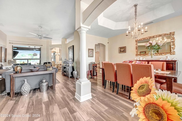 dining area with ceiling fan with notable chandelier, a raised ceiling, wood-type flooring, and ornate columns