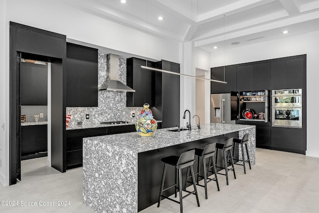 kitchen with stainless steel appliances, decorative backsplash, wall chimney exhaust hood, sink, and an island with sink