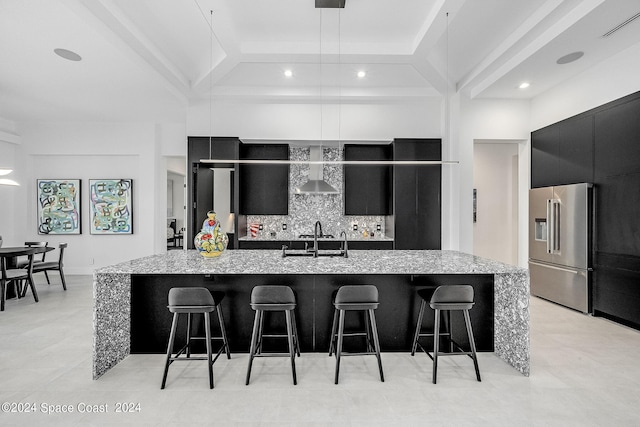 kitchen featuring decorative light fixtures, a large island with sink, sink, wall chimney range hood, and high quality fridge