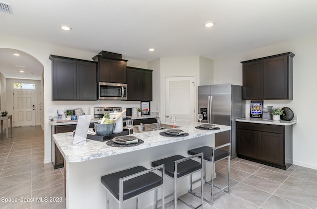 kitchen with appliances with stainless steel finishes, dark brown cabinetry, a kitchen island with sink, a kitchen breakfast bar, and light tile patterned flooring