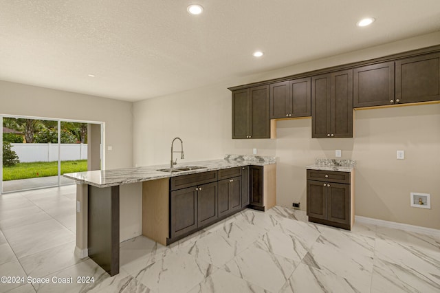 kitchen with dark brown cabinetry, a peninsula, a sink, marble finish floor, and light stone countertops