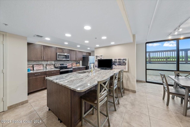 kitchen with appliances with stainless steel finishes, light stone countertops, a kitchen island with sink, tasteful backsplash, and a breakfast bar area