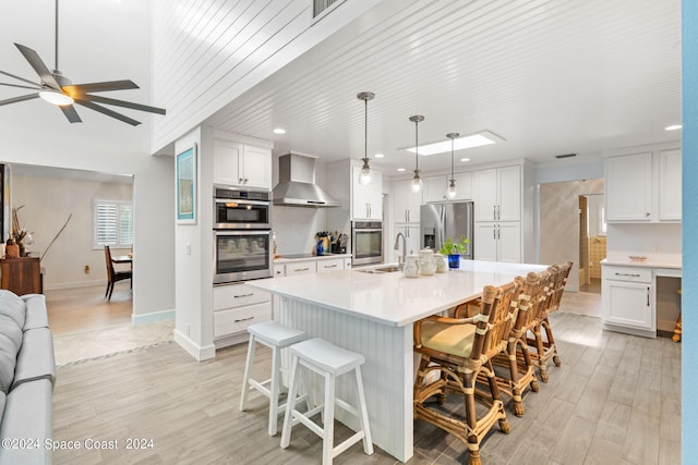 kitchen featuring white cabinets, appliances with stainless steel finishes, light hardwood / wood-style floors, wall chimney range hood, and ceiling fan