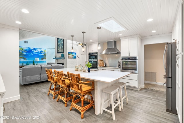 kitchen featuring wall chimney exhaust hood, an island with sink, a kitchen bar, a skylight, and light wood-type flooring