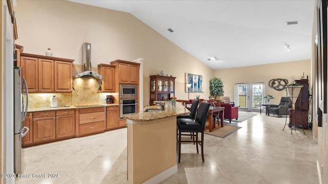 kitchen with appliances with stainless steel finishes, a kitchen island with sink, a breakfast bar, light stone counters, and wall chimney range hood