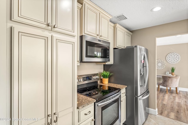 kitchen with cream cabinets, dark stone countertops, a textured ceiling, light hardwood / wood-style floors, and stainless steel appliances