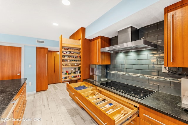 kitchen with wall chimney exhaust hood, light hardwood / wood-style floors, decorative backsplash, black electric stovetop, and dark stone counters