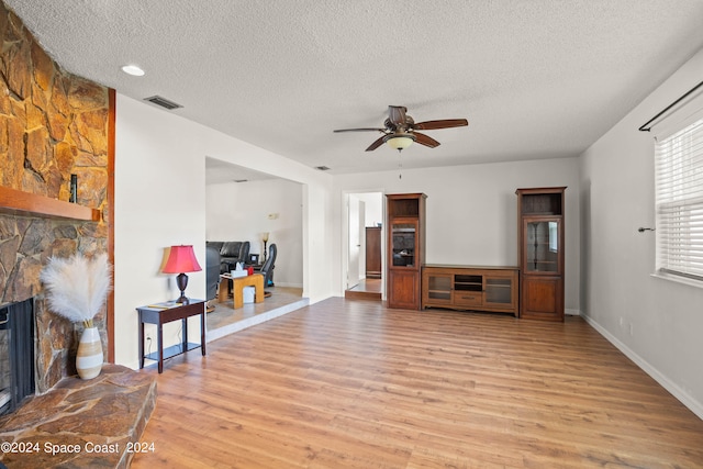 living room with light hardwood / wood-style floors, a textured ceiling, a stone fireplace, and ceiling fan