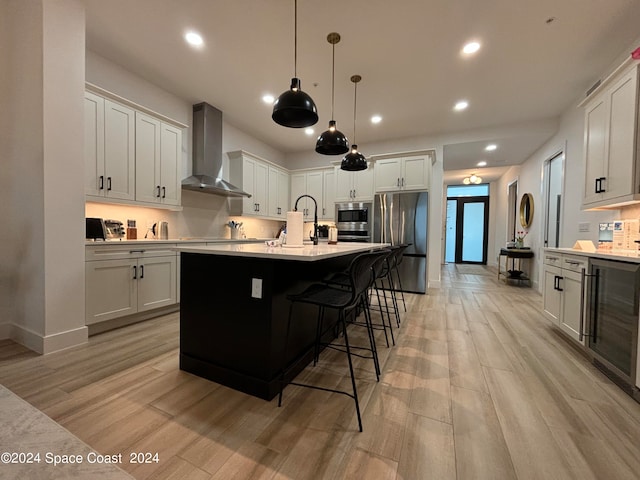 kitchen featuring white cabinetry, wall chimney range hood, an island with sink, appliances with stainless steel finishes, and light hardwood / wood-style floors