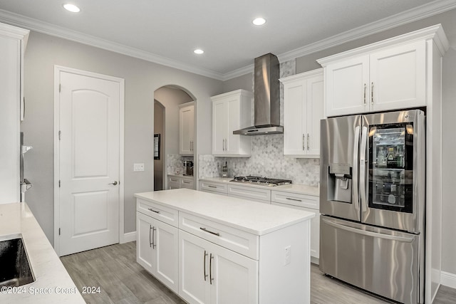 kitchen with white cabinets, ornamental molding, wall chimney range hood, appliances with stainless steel finishes, and light wood-type flooring