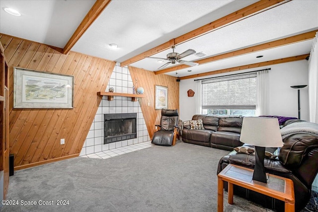 living room featuring wood walls, a tiled fireplace, ceiling fan, and beamed ceiling