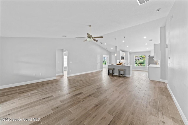 unfurnished living room with arched walkways, a ceiling fan, vaulted ceiling, light wood-type flooring, and baseboards