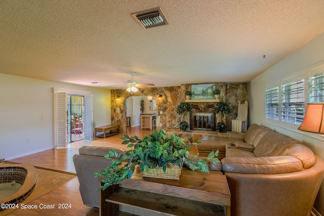 living room featuring ceiling fan, a stone fireplace, a textured ceiling, and light hardwood / wood-style flooring