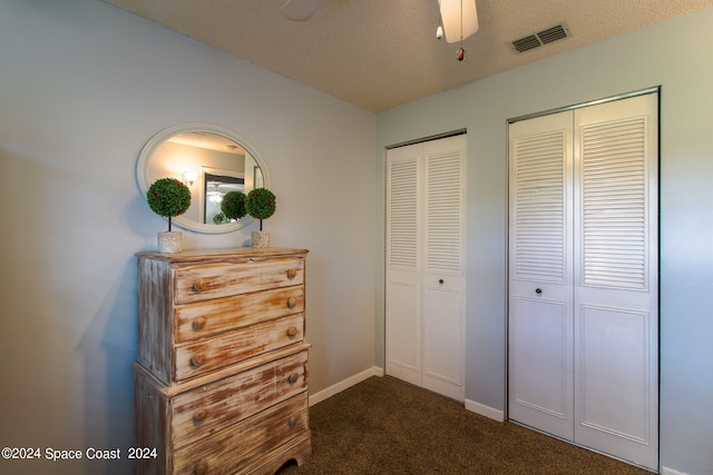 unfurnished bedroom featuring ceiling fan, a textured ceiling, two closets, and dark colored carpet