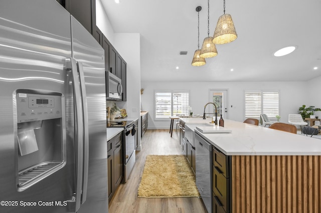 kitchen featuring sink, hanging light fixtures, stainless steel appliances, a center island with sink, and light wood-type flooring