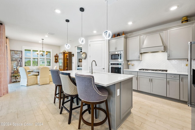 kitchen featuring premium range hood, an island with sink, gray cabinetry, decorative light fixtures, and appliances with stainless steel finishes