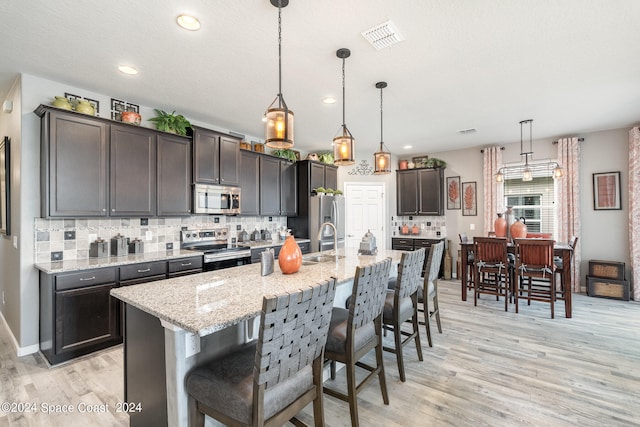 kitchen with an island with sink, stainless steel appliances, hanging light fixtures, and dark brown cabinets