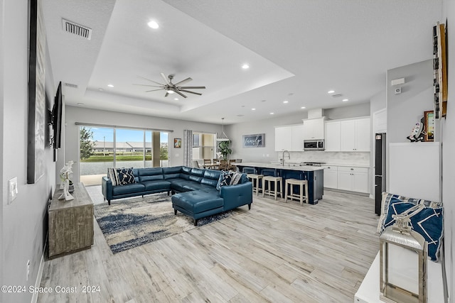 living room featuring light wood-type flooring, a tray ceiling, a textured ceiling, sink, and ceiling fan