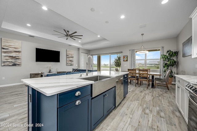 kitchen featuring light wood-type flooring, a tray ceiling, sink, a center island with sink, and blue cabinetry