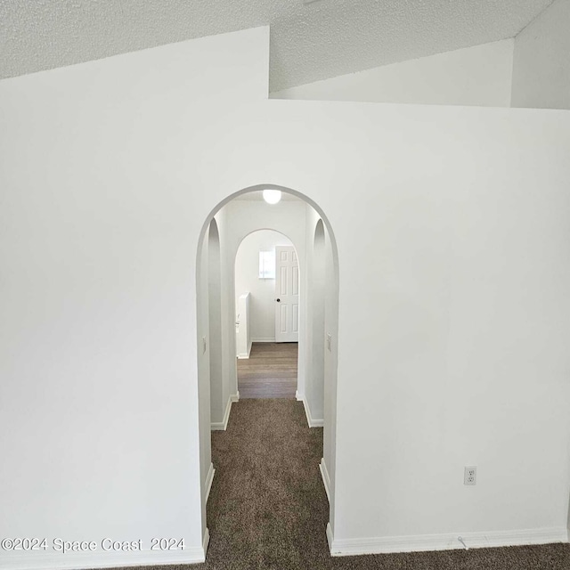 corridor with a textured ceiling, vaulted ceiling, and dark colored carpet