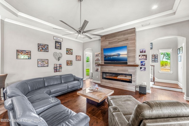 living room with ceiling fan, a raised ceiling, dark hardwood / wood-style floors, a tiled fireplace, and ornamental molding