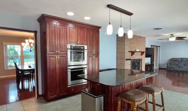 kitchen featuring light tile patterned flooring, hanging light fixtures, a brick fireplace, double oven, and ceiling fan with notable chandelier