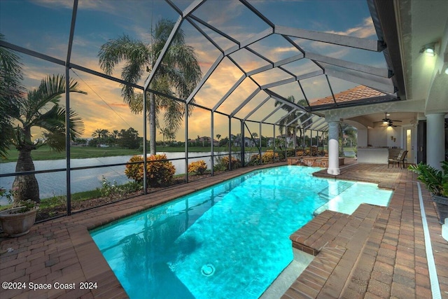 pool at dusk featuring ceiling fan, a patio, glass enclosure, and a water view