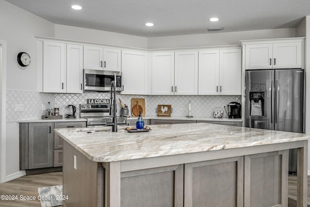 kitchen featuring light wood-type flooring, light stone countertops, an island with sink, white cabinets, and appliances with stainless steel finishes