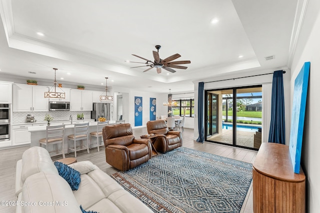 living room with ceiling fan with notable chandelier, light wood-type flooring, crown molding, and a tray ceiling