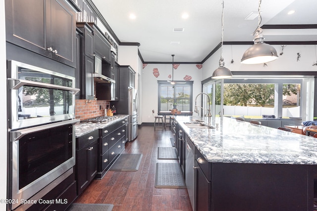 kitchen with a large island with sink, hanging light fixtures, ornamental molding, appliances with stainless steel finishes, and dark hardwood / wood-style flooring