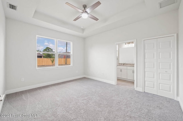 unfurnished bedroom with light carpet, ensuite bathroom, a tray ceiling, ceiling fan, and a closet