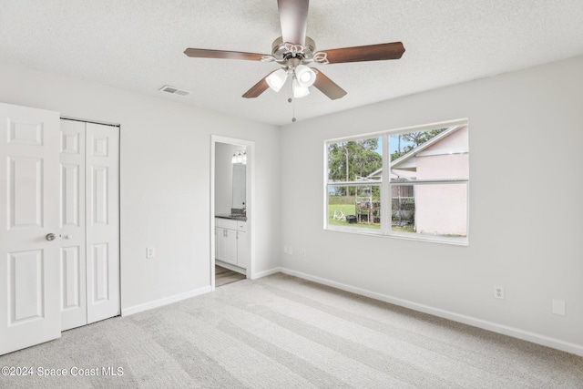 unfurnished bedroom featuring a closet, ensuite bath, light colored carpet, a textured ceiling, and ceiling fan