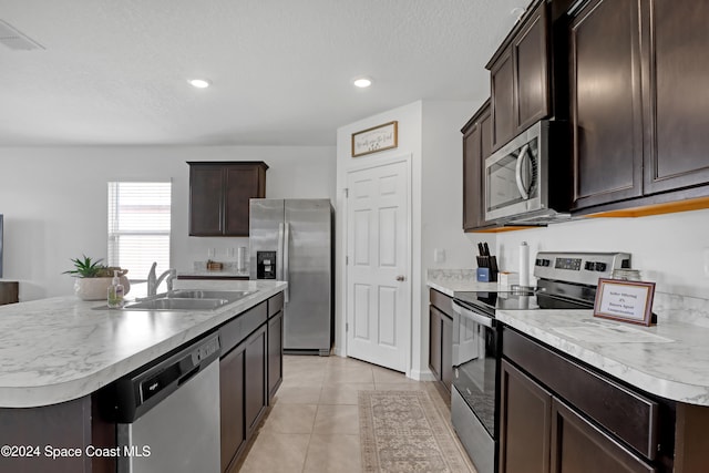 kitchen with stainless steel appliances, dark brown cabinetry, light tile patterned floors, sink, and an island with sink