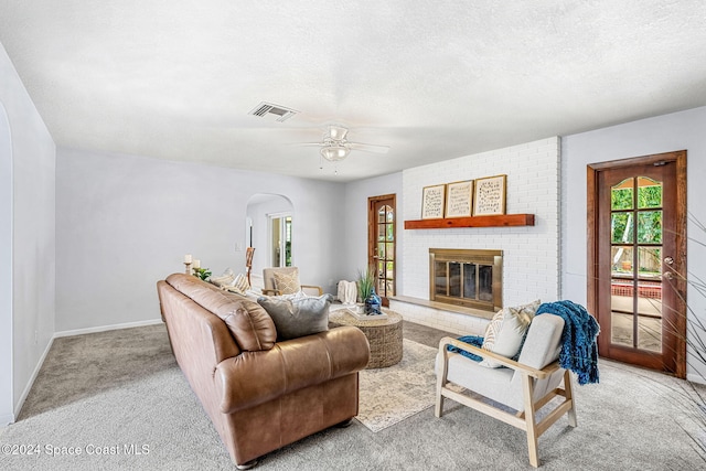 living room featuring ceiling fan, a textured ceiling, a brick fireplace, and light colored carpet