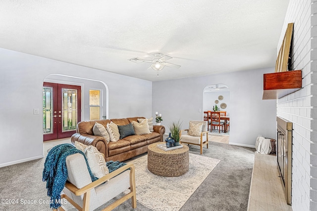 living room with french doors, a textured ceiling, a brick fireplace, ceiling fan, and light colored carpet