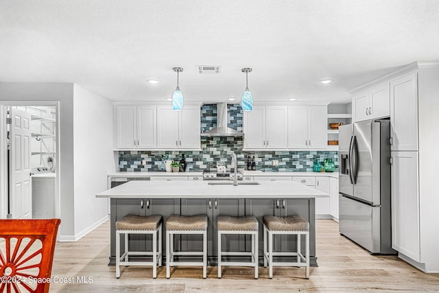 kitchen featuring wall chimney range hood, white cabinets, a kitchen island with sink, light hardwood / wood-style flooring, and stainless steel refrigerator with ice dispenser