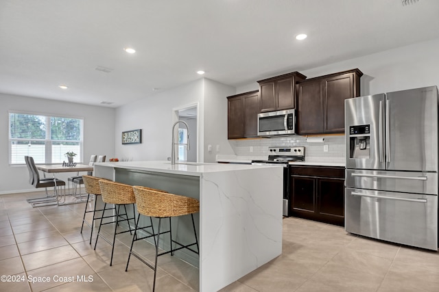 kitchen featuring an island with sink, stainless steel appliances, backsplash, dark brown cabinetry, and light tile patterned floors