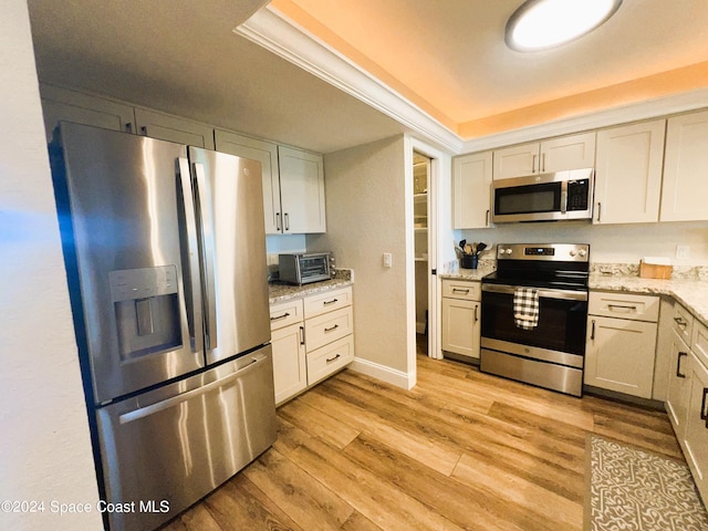 kitchen with appliances with stainless steel finishes, light wood-type flooring, a tray ceiling, white cabinets, and light stone counters