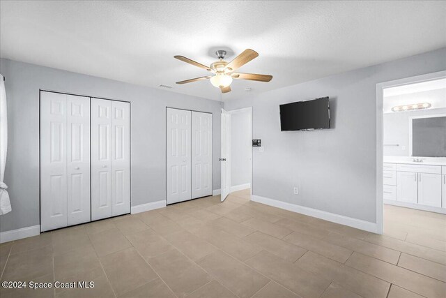 unfurnished bedroom with ensuite bath, a textured ceiling, ceiling fan, light tile patterned floors, and two closets